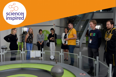 Osiem osób stoi dookoła eksponatu z wahadłem, w rogu zdjęcia logo Science Inspired