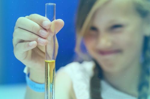 Na pierwszym planie zlewka chemiczna z płynem, na drugim planie uśmiechnięta twarz dziewczyny