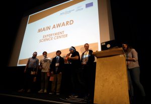 Sześć osób stoi na scenie, jedna z nich mówi przez mikrofon. W tle slajd z napisem Main Award EXPERYMENT Science Center