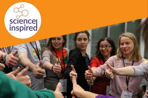 Grupa osób stoi w kółku, pokazując znak ok, w rogu zdjęcia logo Science Inspired