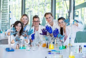 dla grup kafel Trzy dziewczyny i dwóch chłopaków pozuje do zdjęcia w niebieskich rękawiczkach, wszyscy się uśmiechają. Przed nimi na stole leżą liczne odczynniki chemiczne i szkło laboratoryjne
