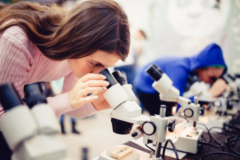 Kobieta spogląda przez mikroskop, w tle jeszcze jedna osoba używająca mikroskopu