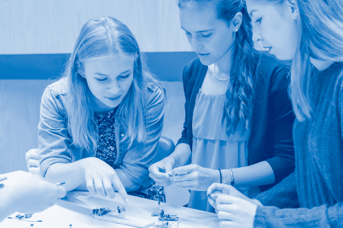 Niebieskie zdjęcie trzech nastolatek pochylających się nad stołem z małymi robotami.