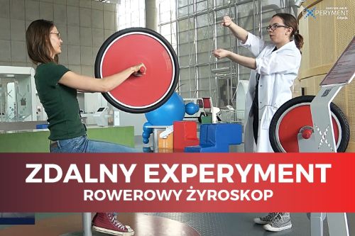 Zdalny experyment, rowerowy żyroskop. Jedna edukatorka siedzi trzymając w dłoniach duże czerwone koło, druga tłumaczy jej działanie żyroskopu.