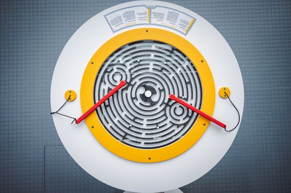 Widok z góry na labirynt w kształcie koła. Wewnątrz labiryntu mała metalowa kulka. Obok leżą dwie czerwone pałeczki.