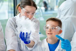 warsztaty-dla-rodzin-EXPERYMENT Kobieta w okularach ochronnych prezentuje doświadczenie chemiczne chłopcu w okularach ochronnych