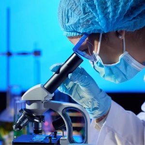 Kobieta ubrana w fartuch laboratoryjny, maseczkę, czepek, okulary i rękawiczki patrzy przez mikroskop.