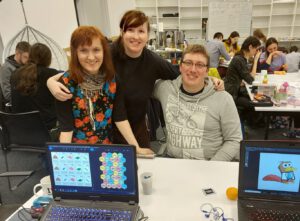 1 Na fotografii uwiecznione zostało kilkanaście osób, biorących udział w wydarzeniu naukowym. W centrum forografii widać dwie kobiety oraz meżczyzne, wszystkie trzy osoby pozują oraz uśmiechają się do zdjęcia. W tle za nimi na białej tablicy widać tytuł warsztatów który brzmi "Space Game Jam 2020", a pod napisem godzinową rozpiskę wydarzenia.