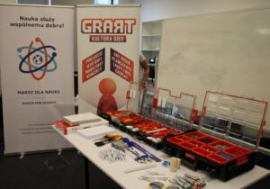 244511580_988502991727312_5162771276363081232_n Na zdjęciu mamy stól, na którym umieszczone są 3 organizery, w których znajdują się różnego rodzaju znaczniki oraz pionki. Na stole leżą również linijki, długopisy, markety, żetony, gumki, nożyczki, taśmy i ryza papieru A4. W tle, za stołem stoją dwa banery, promujące wydarzenia "Marsz Dla Nauki" i "GRART Klutura Gier"