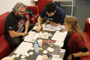 244621371_988499788394299_7729469942889927186_n Zdjęcie przedstawia trzy osoby siedzące na czerwonych kanapach, wokół czarnego stołu. Na stole znajduje się mnóstwo rozpisanych kartek, a w centrum stołu leży coś na wzór gry planszowej, składającej się z położonych obok siebie sześciokątów, na których umieszczone są kolorowe kwadraciki. Jedna z osób właśnie podnosi kwadracik i pradopodobnie będzie go umieszczać na którymś z sześciokątów.