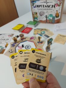 Impulsacja Fotografia przedstawia grę planszową pt. "Impulsacja: Ostatnia Szara Komórka". Na głównym planie zdjęcia osoba trzyma w dłoni trzy karty, a jedna z nich zatytułowana jest "Liście Manayupa", mamy również informacje jakie funkcje ma ta karta w grze. W tle zdjęcia widać plansze gry, na której znajdują się pionki, a wokół planszy rozłożone są inne karty w żółtym, czerwonym i zielonym kolorze.