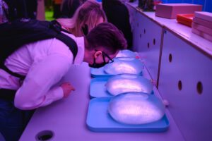 Zdjęcie przedstawia młodą parę osób, mężczyzna ubrany jest w białą koszulę i ma na sobie plecak oraz okulary, a kobieta za nim ubrała się w kolorową sukienke. Obie osoby pochylone są nad stołem i przyglądają się prezerwatywom, wypełnionym cieczą, które leżą na niebieskich podstawkach.