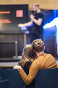 Zdjęcie przedstawia parę siędzącą bardzo blisko siebie podczas pokazu, prowadzonego przez jednego z edukatorów CN Experyment. Para styka się bokami głów, a Pan trzyma swoją lewą ręke na krześle partnerki, za jej plecami.