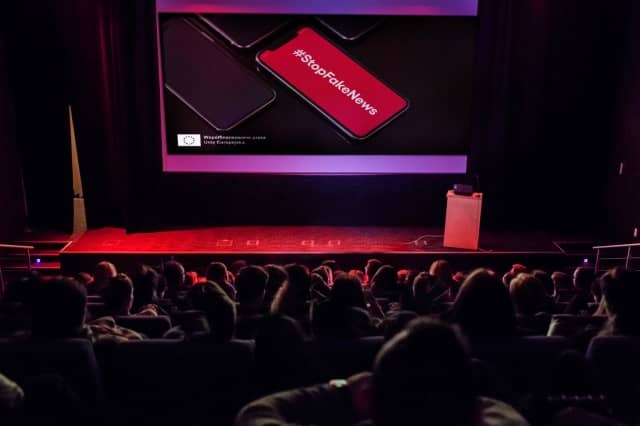 Sala wykładowa pełna ludzi, na ekranie prezentacja z wyświetlonym na czerwonym tle napisem #StopFakeNews.