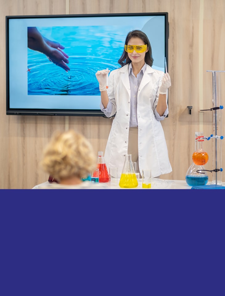 Kobieta w fartuchu laboratoryjnym i okularach stoi za stołem z kolorowymi cieczami i sprzętem laboratoryjnym. W tle ekran z wodą.