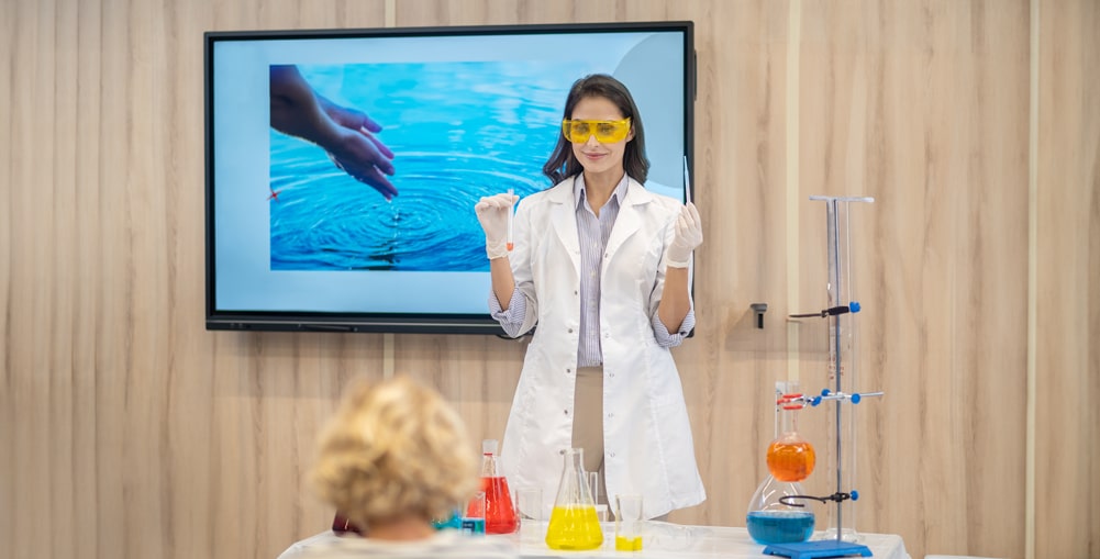 Kobieta w fartuchu laboratoryjnym i okularach stoi za stołem z kolorowymi cieczami i sprzętem laboratoryjnym. W tle ekran z wodą.