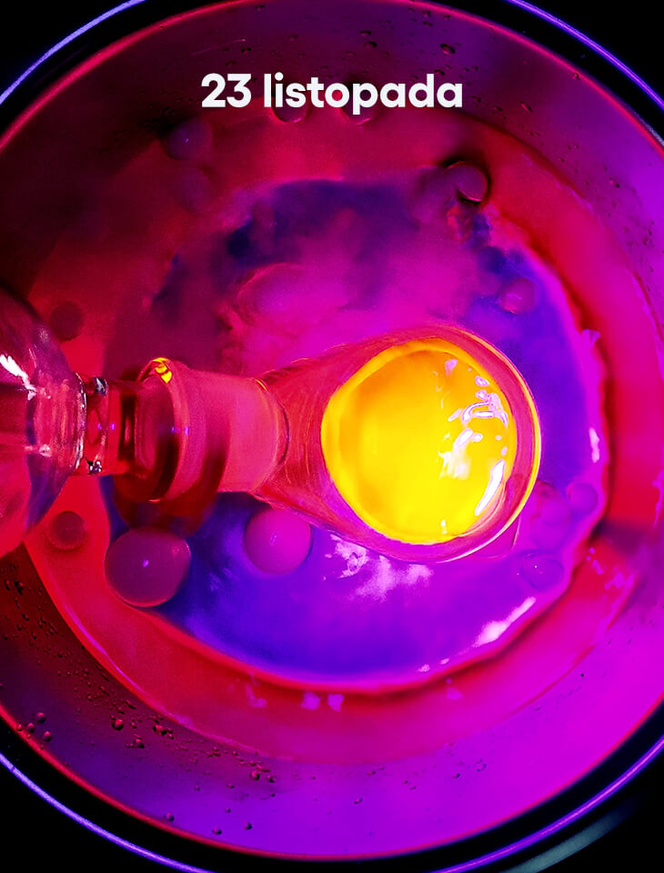Kolorowa scena laboratoryjna ukazana z góry: w różowo-fioletowym świetle widać naczynie z fluorescencyjną cieczą i żółtą, świecącą kroplą, u góry biały napis ‘23 listopada’