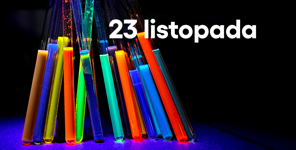 Kilka probówek z kolorowymi, fluorescencyjnymi płynami ustawionych w półokręgu na ciemnym tle, z białym napisem ‘23 listopada’ u góry.