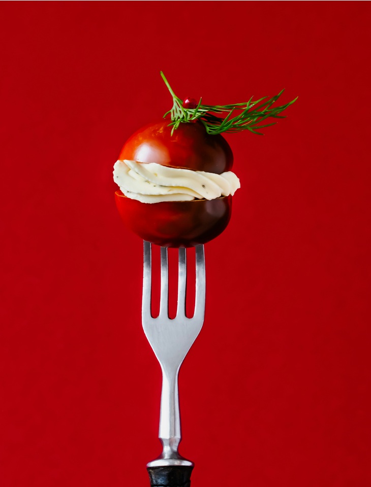 Na widelcu nabity jest przekrojony na pół czerwony pomidor, przełożony grubym plasterkiem białego sera. Na górze leży gałązka zielonego koperku. Całość sfotografowana na intensywnie czerwonym tle, w minimalistycznym, studyjnym stylu.